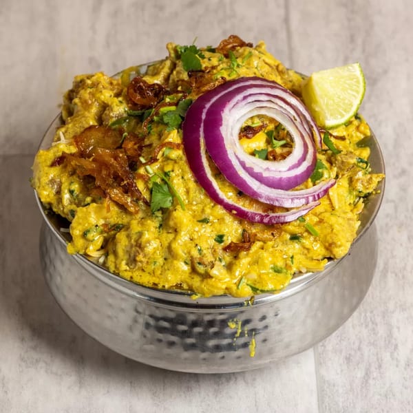 Pista House's creamy yellow Haleem in a silver bowl, garnished with fried onions, red onion, and lime.